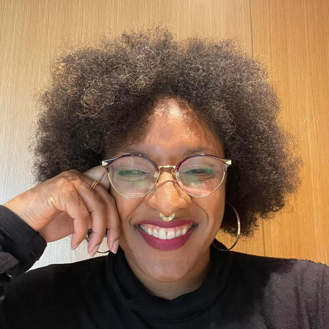 Joya Jones, a radiant Black woman with a voluminous afro, wears stylish gold-rimmed glasses, a septum ring, and a black turtleneck. Resting her head on her hand, she smiles confidently, exuding intelligence, creativity, and effortless charm.